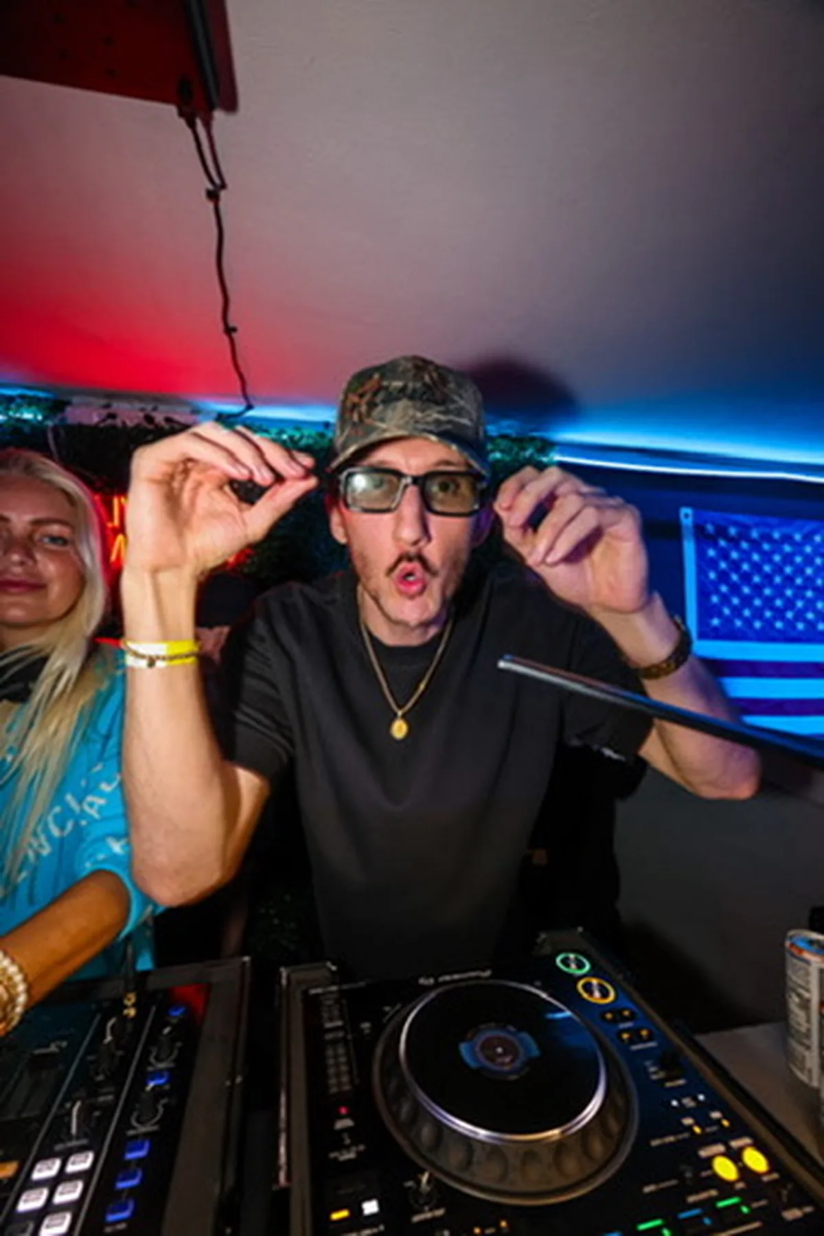 DJ adjusting sunglasses mid-set with neon lighting behind him