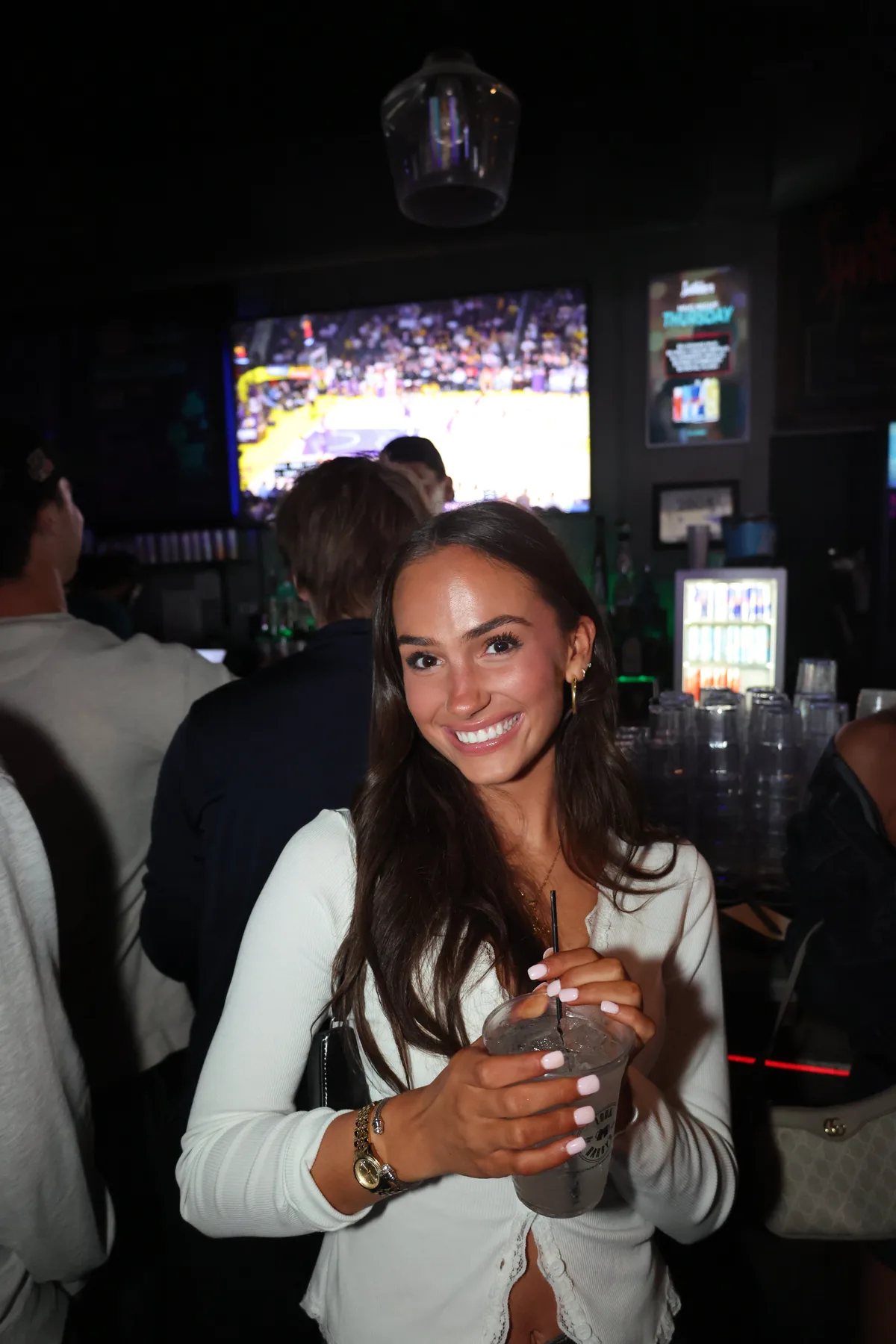 Woman holding a drink at the bar with a game on screen behind her